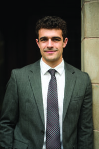 Anthony Potts, wearing a gray pinstripe suit and patterned tie, smiles for a portrait while leaning against a stone doorway.