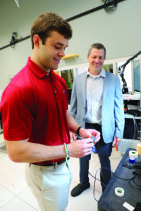 Carson Kennedy holds a small sensor device while Dr. Christopher Modlesky looks on in a research lab.