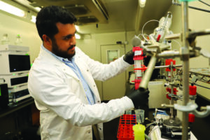 Hanif Zaman, wearing a lab coat and gloves, adjusts a glass column on lab equipment in a biochemistry lab.