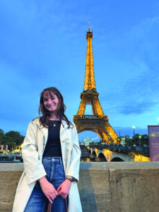 Jacqueline GaNun smiles in front of the illuminated Eiffel Tower at dusk, with the Seine River and a bridge visible behind her.